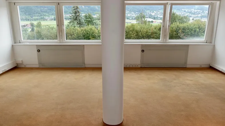 Geräumiger Büroraum mit Teppichboden, Säule und großen Fenstern mit Blick ins Grüne.