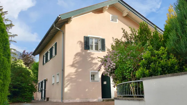 Traditionelles Wohnhaus mit beigefarbener Fassade, grünen Fensterläden und gepflegtem Gartenbereich.
