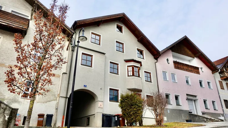 Ansicht eines Mehrfamilienhauses mit heller Fassade, braunen Fenstern und einem Torbogen.
