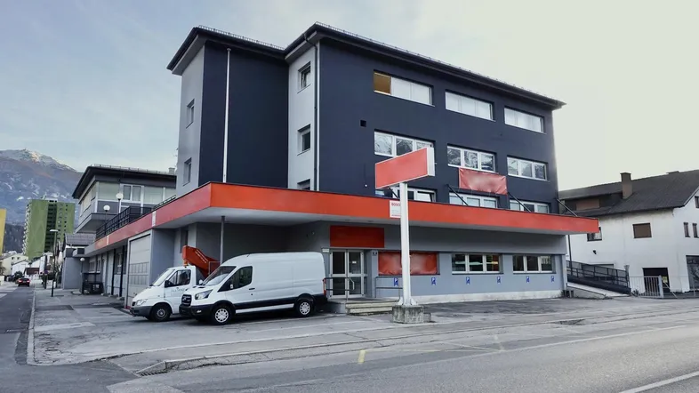 Modernes Geschäftsgebäude mit dunkler Fassade und roten Akzenten, zwei Lieferwagen parken davor.