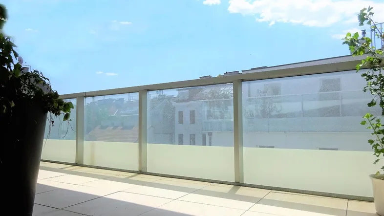 Sonnige Terrasse mit modernen Fliesen, Glasgeländer und Blick auf die umliegenden Gebäude.