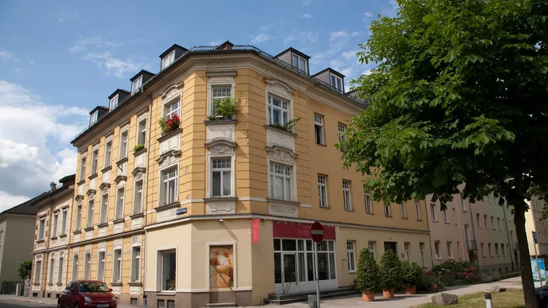 Historisches Eckgebäude mit gelber Fassade und Geschäftsräumen im Erdgeschoss, umgeben von Bäumen und Straße.