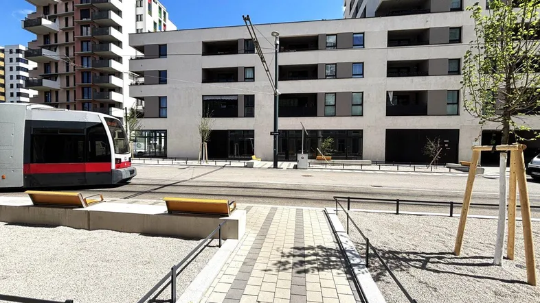 Moderne Gebäude entlang einer Straße mit Straßenbahn und neu angelegten Gehwegen unter blauem Himmel.