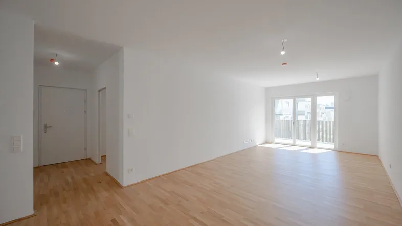 Helles, leeres Zimmer mit Parkettboden und Zugang zu einem Balkon oder einer Terrasse.
