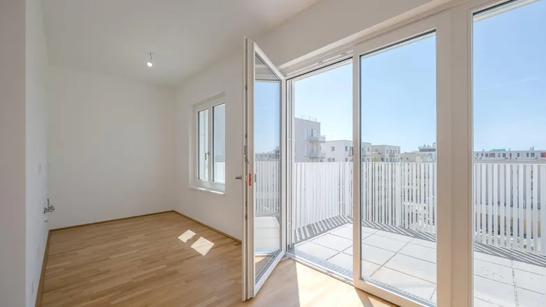 Helles, leeres Zimmer mit Holzboden und großem Zugang zu einer sonnigen Terrasse.