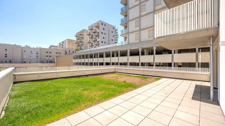 Große geflieste Terrasse mit Grünfläche und Blick auf moderne Wohngebäude.