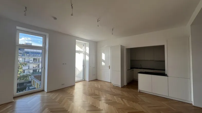 Heller Wohnbereich mit integrierter Küche, Fischgrätparkett und Zugang zu einem Balkon.
