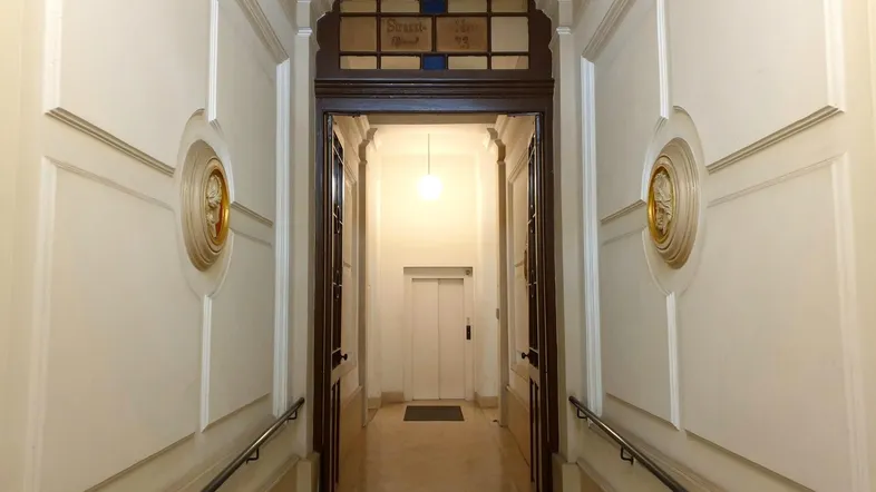Langer Flur mit dekorativen Wandelementen und Zugang zu einem Aufzug im Altbau.