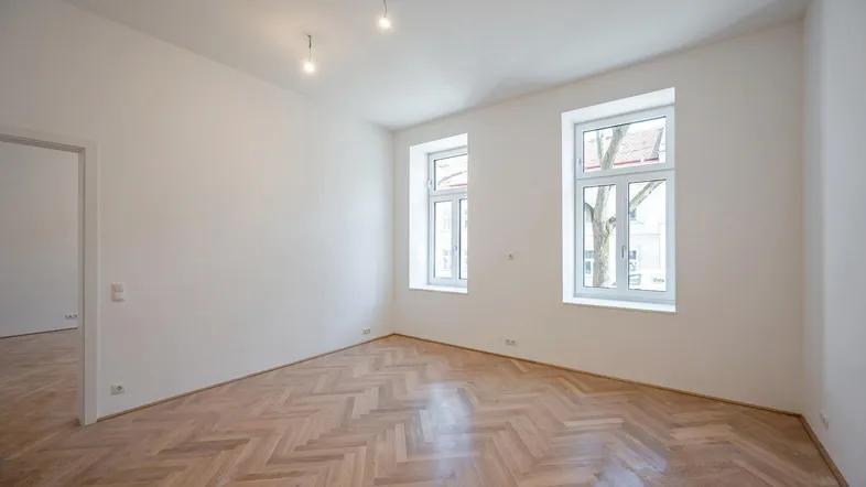 Heller, leerer Raum mit Fischgrätparkett und zwei großen Fenstern, die viel Tageslicht hereinlassen.