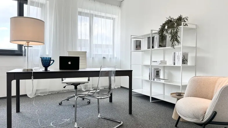 Modernes Büro mit Schreibtisch, transparentem Stuhl und weißem Regal, gut beleuchtet durch große Fenster.