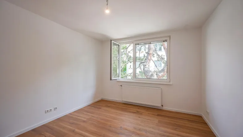 Leeres Zimmer mit Holzboden und einem geöffneten Fenster, das viel Tageslicht hereinlässt.