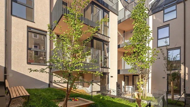 Gemeinschaftlicher Gartenbereich mit Sitzbank, Sandkasten und Blick auf die Balkone des Neubaus.