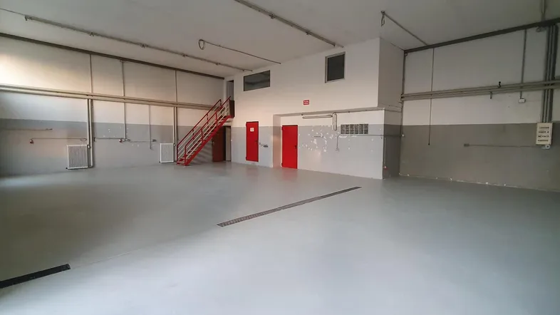 Geräumige Werkshalle mit hohen Decken, hellgrauem Boden und einer roten Treppe im Hintergrund.