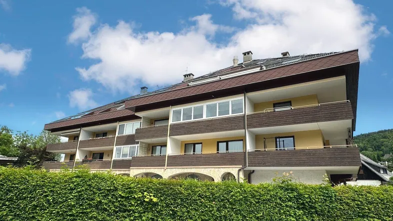 Mehrfamilienhaus mit mehreren Balkonen und gepflegter Hecke im Vordergrund unter blauem Himmel.