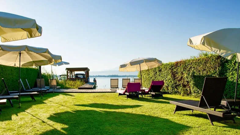 Großer Rasenbereich mit Liegestühlen und Sonnenschirmen direkt am Seeufer mit Steg.