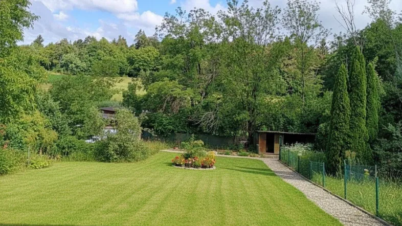 Gepflegter Rasenbereich mit Blumenbeet und Gartenhaus, umgeben von üppiger Vegetation und Bäumen.