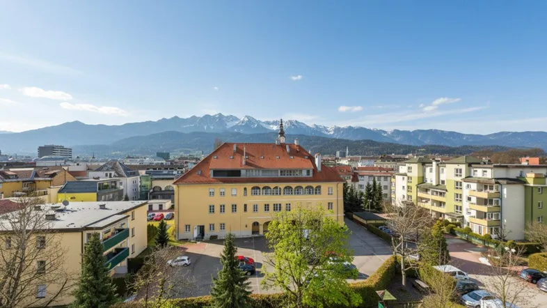 Panoramablick über die Stadt mit Gebäuden, Grünflächen und majestätischen Bergen im Hintergrund.