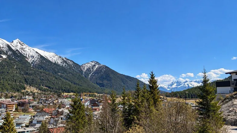 Panoramablick auf schneebedeckte Berge und ein Dorf in einem Tal unter blauem Himmel.