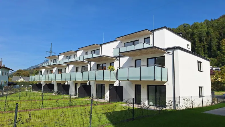 Moderne Reihenhäuser mit weißen Fassaden, Balkonen und kleinen Gärten vor einer grünen Bergkulisse.