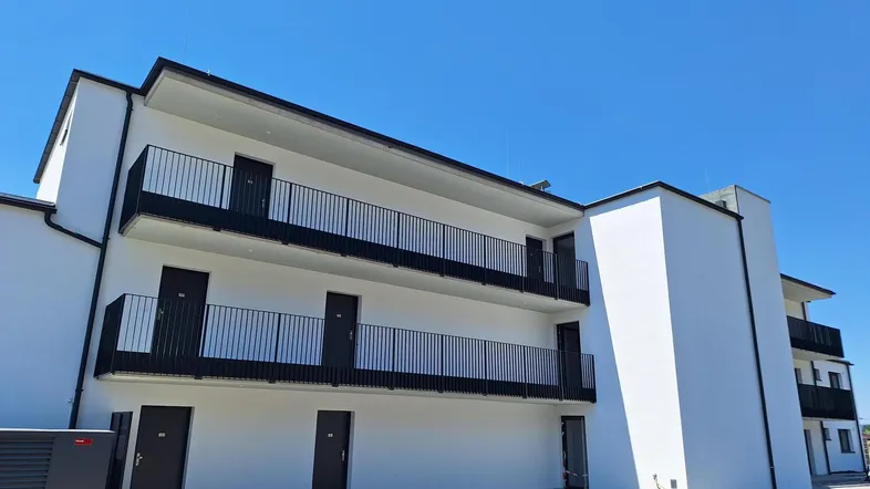 Modernes Mehrfamilienhaus mit weißer Fassade, schwarzen Balkonen und Flachdach unter blauem Himmel.