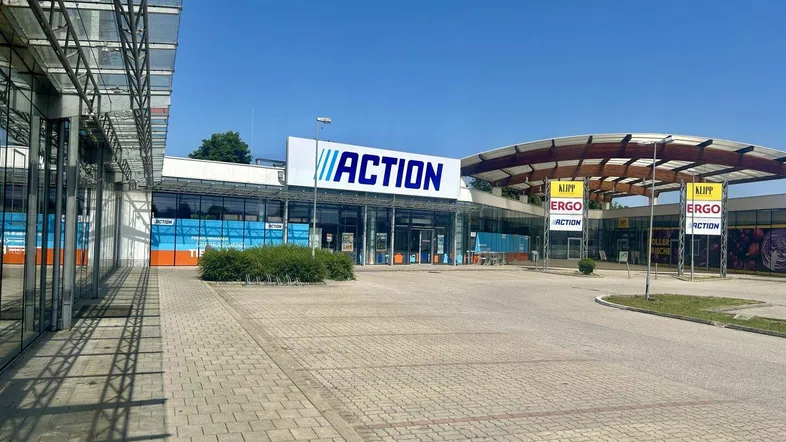 Blick auf ein modernes Einkaufszentrum mit großflächigen Glasfassaden und einem großen Parkplatz unter blauem Himmel.