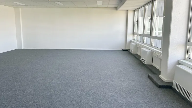Geräumiges, leeres Büro mit grauem Teppichboden und großen Fenstern, die viel Tageslicht hereinlassen.