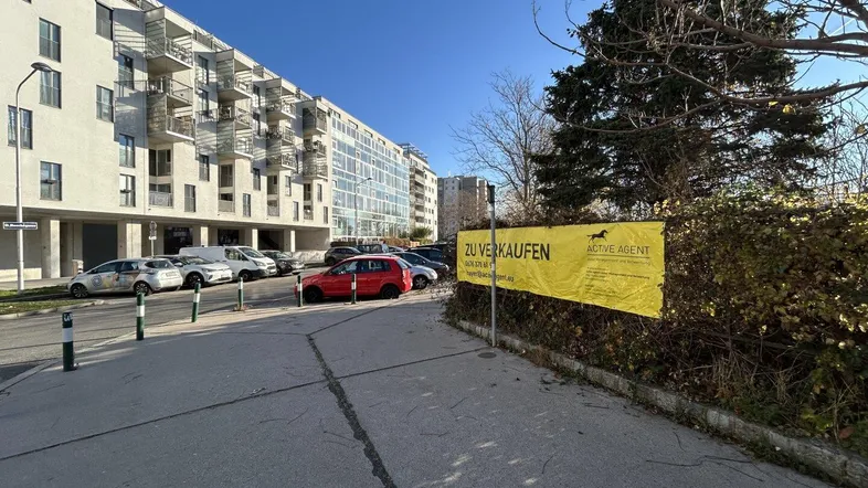 Blick auf eine Straße mit geparkten Autos, modernen Gebäuden im Hintergrund und einem Verkaufsbanner.