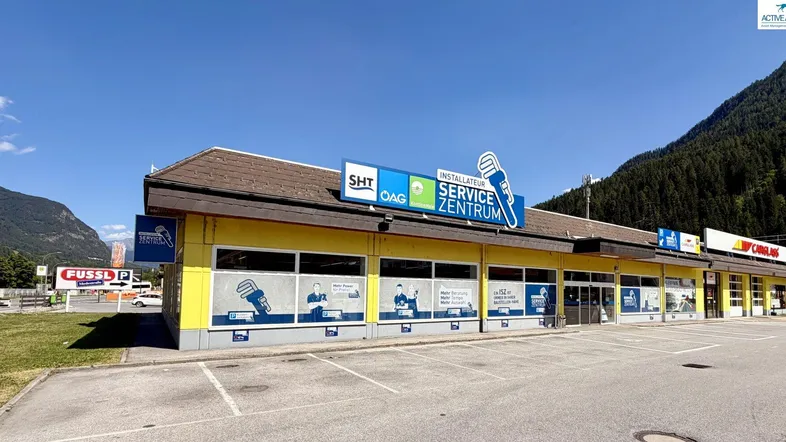Gelbes Geschäftsgebäude mit braunem Dach und großem Parkplatz vor einer beeindruckenden Bergkulisse.