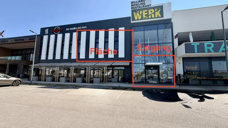 Moderne Geschäftsfassade mit großen Glasflächen und Eingangsbereich, umgeben von einem Parkplatz unter blauem Himmel.