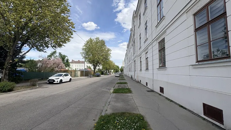 Blick auf die Straße mit einem langen weißen Gebäude und Bäumen unter blauem Himmel.