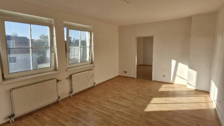 Helles Zimmer mit Holzboden, weißen Wänden und zwei Fenstern, durch die Sonnenlicht einfällt.