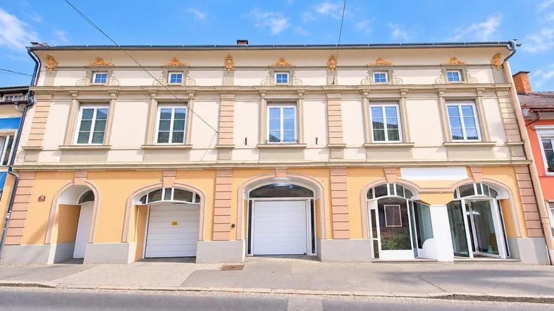 Historisches Gebäude mit einer hellen Fassade, großen Fenstern und drei Garagentoren im Erdgeschoss.