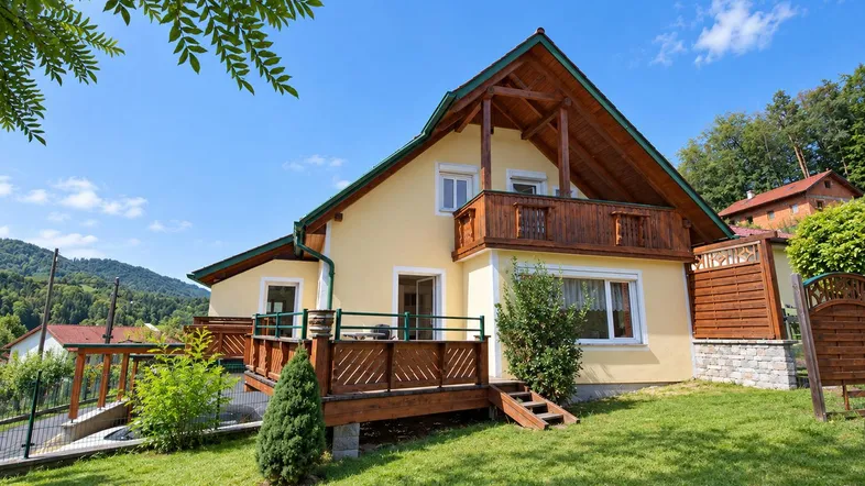 Charmantes Haus mit gelber Fassade, Holzbalkon und großer Holzterrasse im Grünen.