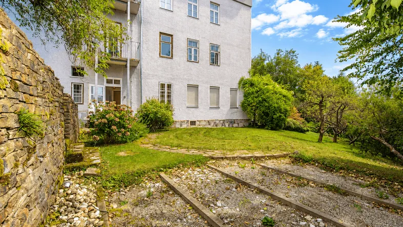 Ansicht des Gebäudes von der Gartenseite mit alter Steinmauer und grüner Rasenfläche unter blauem Himmel.