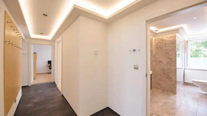 Heller Flur mit moderner indirekter Beleuchtung, dunklem Fliesenboden und Garderobe, führt zu weiteren Räumen.