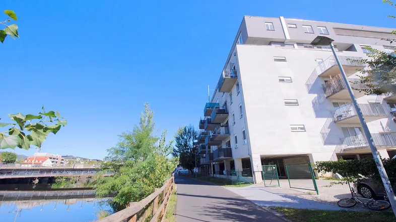 Modernes Mehrfamilienhaus am Flussufer mit Balkonen und Fußweg unter blauem Himmel.