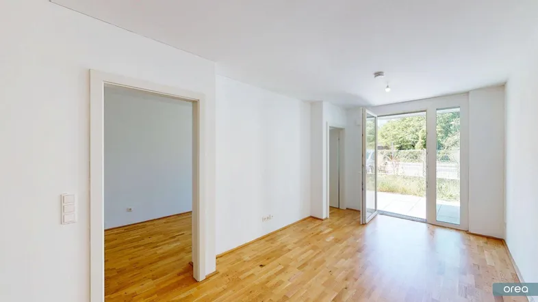 Helles, leeres Wohnzimmer mit Holzboden und Zugang zu einer Terrasse oder einem Gartenbereich.