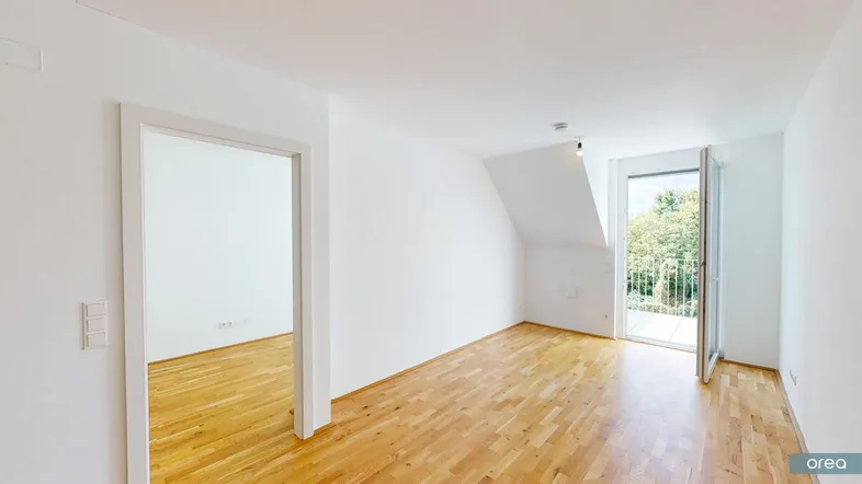Helles Zimmer mit Holzboden, weiß gestrichenen Wänden und Zugang zu einem kleinen Balkon mit Blick ins Grüne.