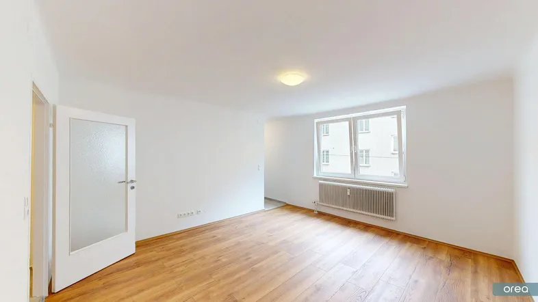 Helles Zimmer mit Laminatboden, weiß gestrichenen Wänden und einem Fenster mit Blick nach draußen.