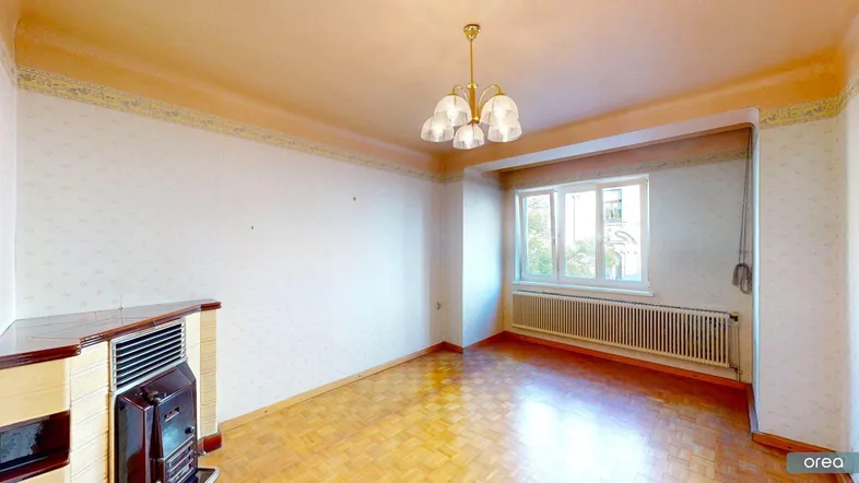 Geräumiges Wohnzimmer mit Fischgrätparkett, Fenster und einem traditionellen Kaminofen, bereit zur Renovierung.