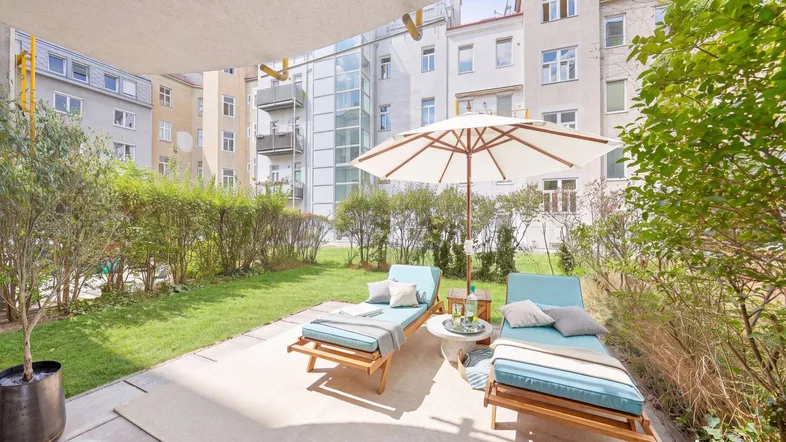Sonniger Gartenbereich mit Liegestühlen, Sonnenschirm und Blick auf die umliegenden Gebäude.