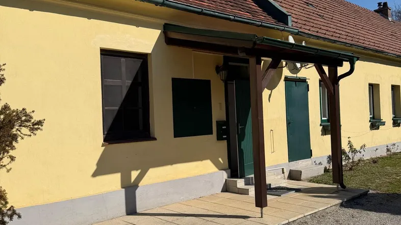 Gelbe Hausfassade mit rotem Ziegeldach, überdachtem Eingangsbereich und gepflastertem Vorplatz.
