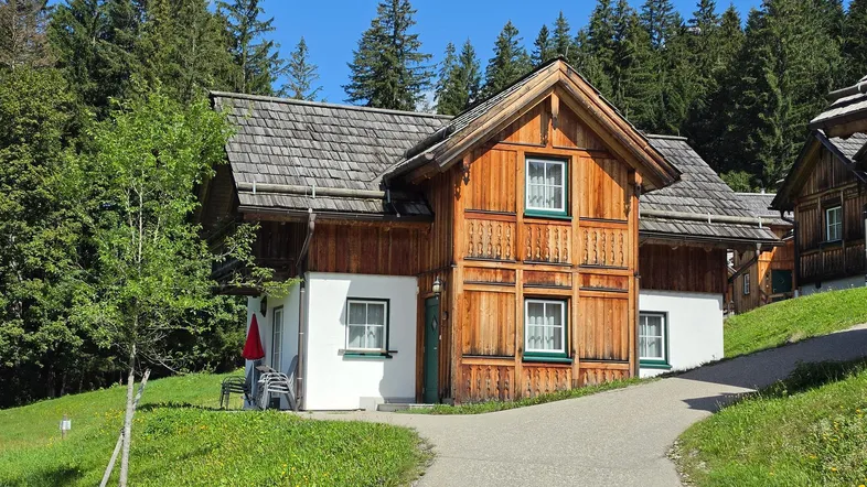 Traditionelles Holzhaus mit weißem Putz und Holzbalkonen, umgeben von grüner Natur und hohen Bäumen.