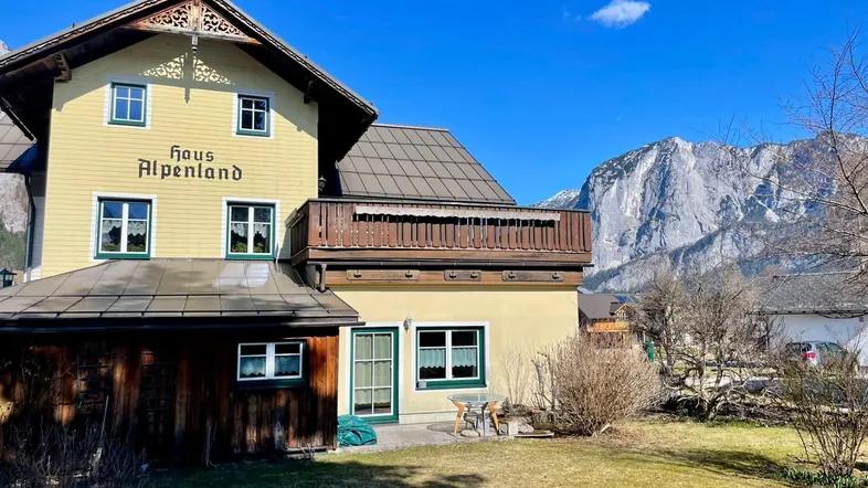 Gelbes Haus im alpinen Stil mit Holzbalkon und beeindruckendem Bergblick unter blauem Himmel.