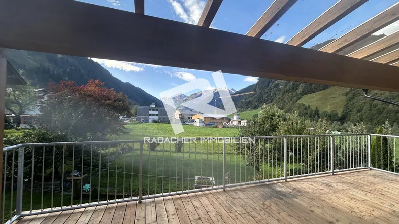 Große Holzterrasse mit Pergola und weitem Blick auf die umliegende Berglandschaft und grüne Wiesen.