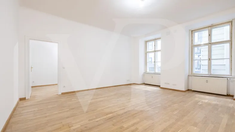 Helles, leeres Zimmer mit Holzboden und zwei großen Fenstern, die viel Tageslicht hereinlassen.