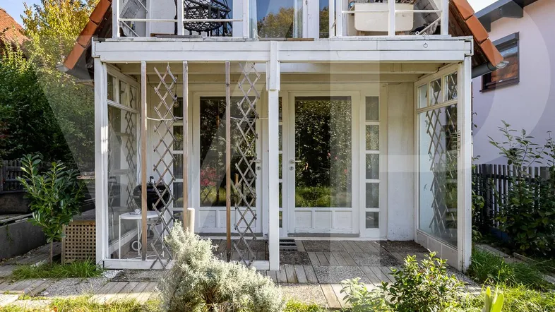 Weißes Haus mit Satteldach, Balkon und Wintergarten, umgeben von einem kleinen Garten.