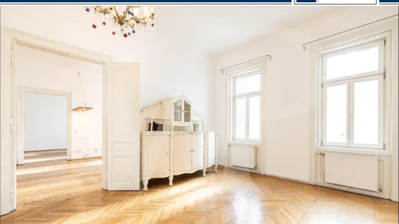 Helles Zimmer mit Fischgrätparkett, hohen Decken und großen Fenstern, verbunden mit einem weiteren Raum.