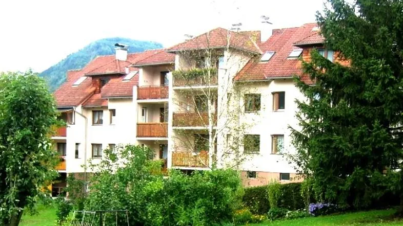 Mehrfamilienhaus mit roten Ziegeldächern, umgeben von viel Grün und Bergen im Hintergrund.