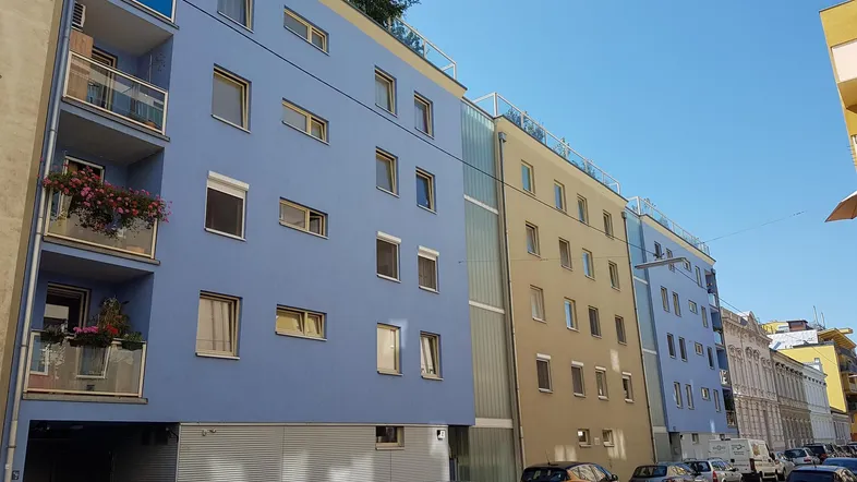 Modernes Wohngebäude mit blauer und beiger Fassade, Balkonen und Dachbegrünung an einer Stadtstraße.